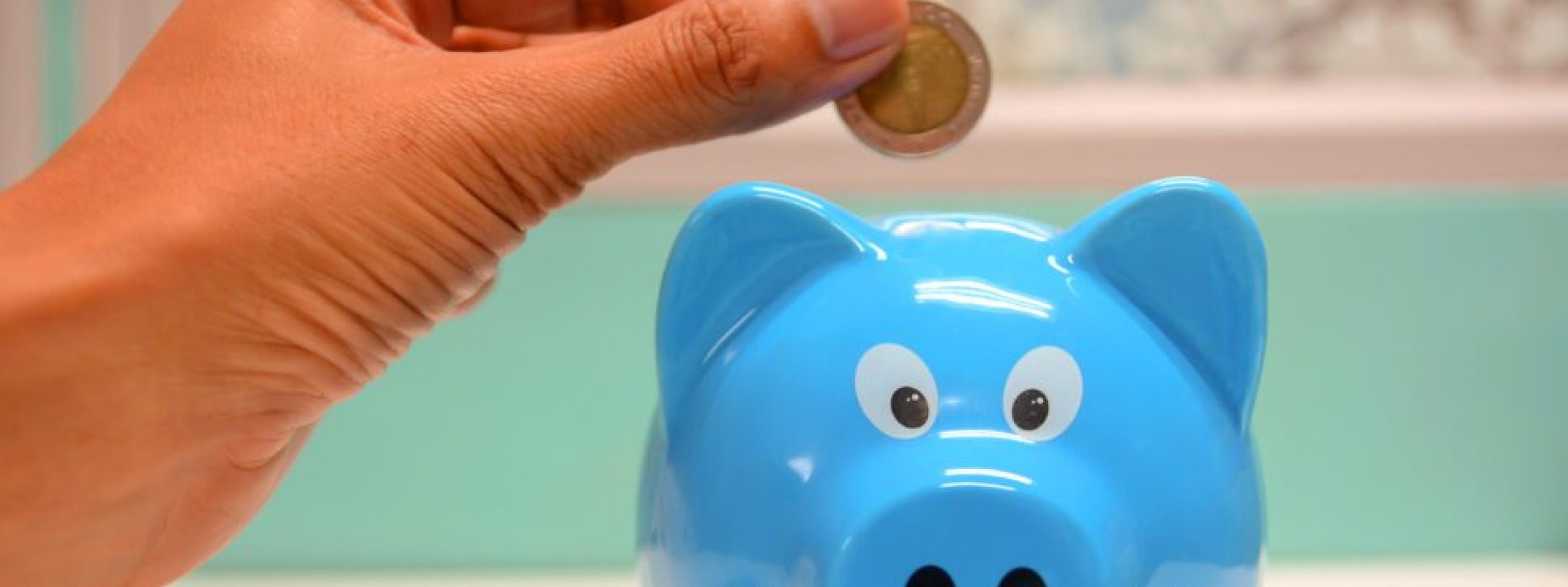 A hand holding a coin over a blue piggy bank.
