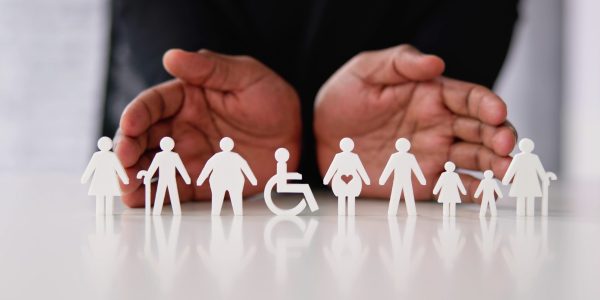 Hands hovering protectively over white paper cut-outs representing diverse individuals, including a pregnant person and someone in a wheelchair