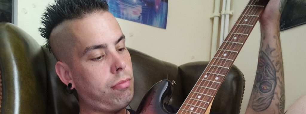 A man with a mohawk hairstyle and tattoos sits in a leather chair, holding an electric guitar