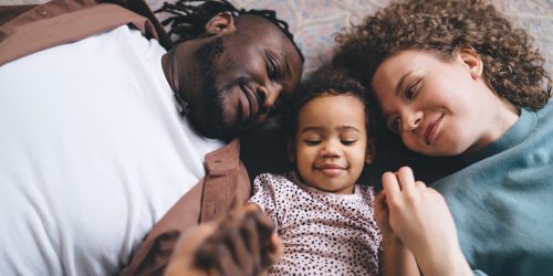 From above of young multiethnic parent in casual clothes with curly hair daughter lying on back over floor holding hands and looking at each other while resting and bonding together in living room