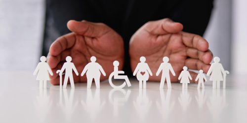 Hands hovering protectively over white paper cut-outs representing diverse individuals, including a pregnant person and someone in a wheelchair