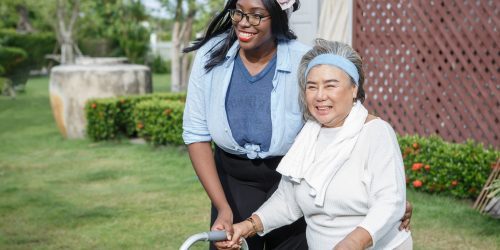 asian senior mother walk with walker and african american carer support in garden at home. old woman disabled walking and black caregiver young woman helping in back yard park outdoors. health care