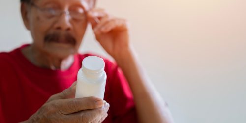 An elderly man in a red shirt holds a white pill bottle while adjusting his glasses