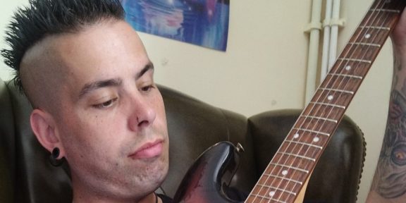 A man with a mohawk hairstyle and tattoos sits in a leather chair, holding an electric guitar